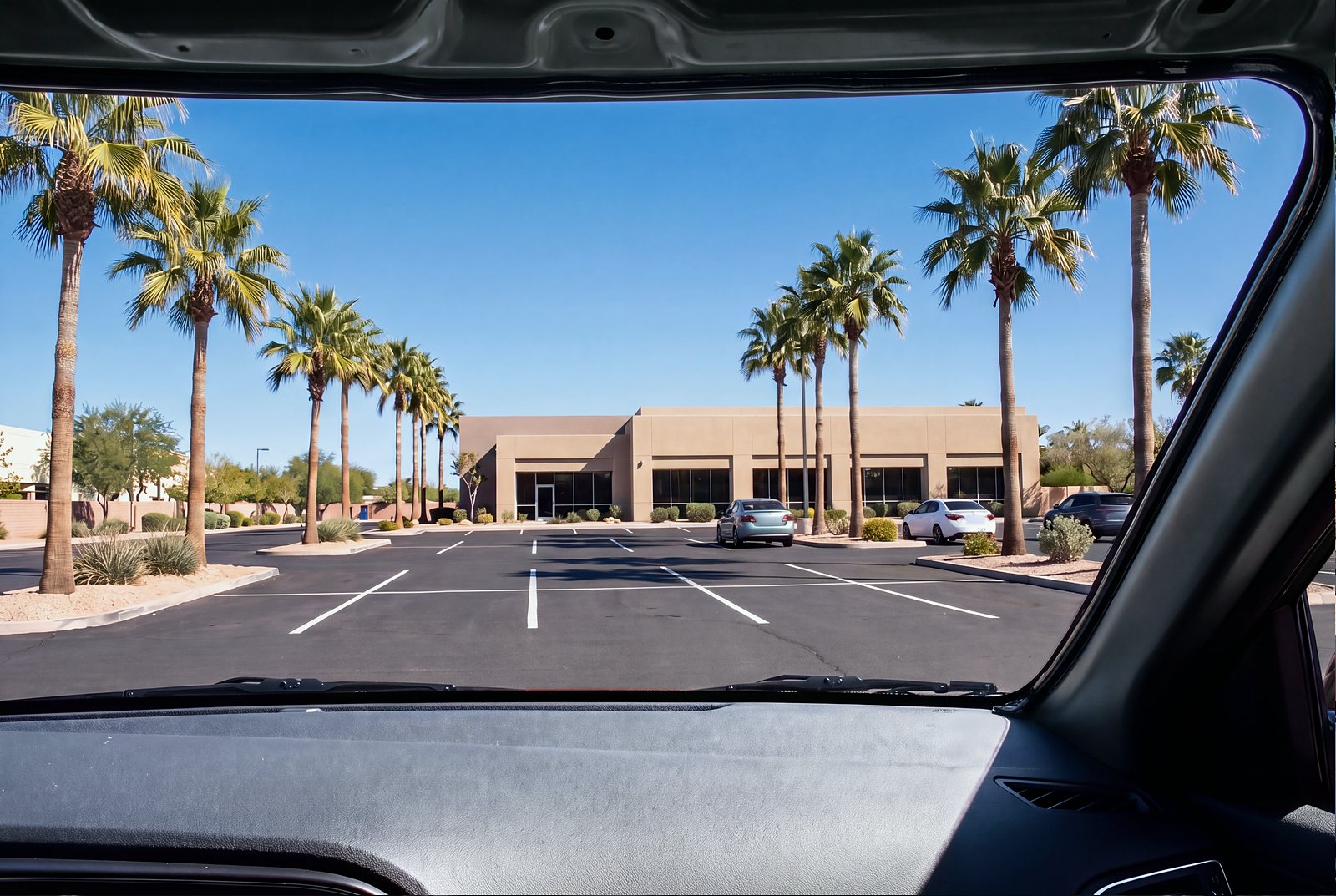 Looking out from the driver's seat through an empty windshield opening, the glass removed and the bare roof edge showing, toward a sunny parking lot.