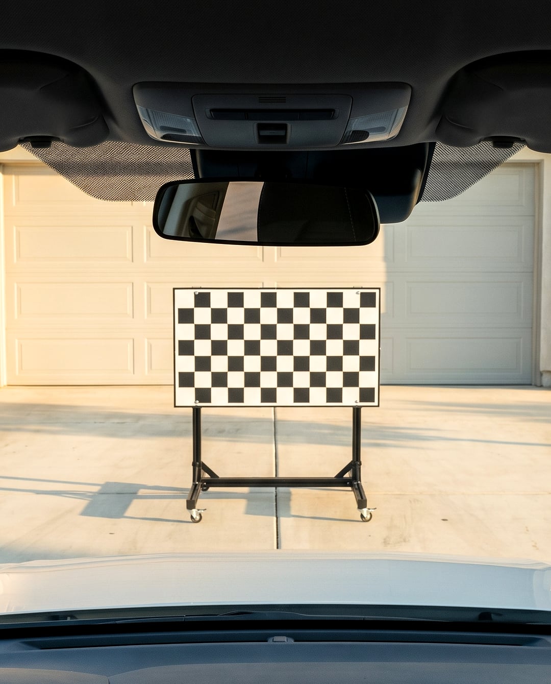 Looking out through a car's windshield, past the rear-view mirror and its mount, at a checkerboard calibration target on a wheeled stand, centred in front of the hood on a home driveway.