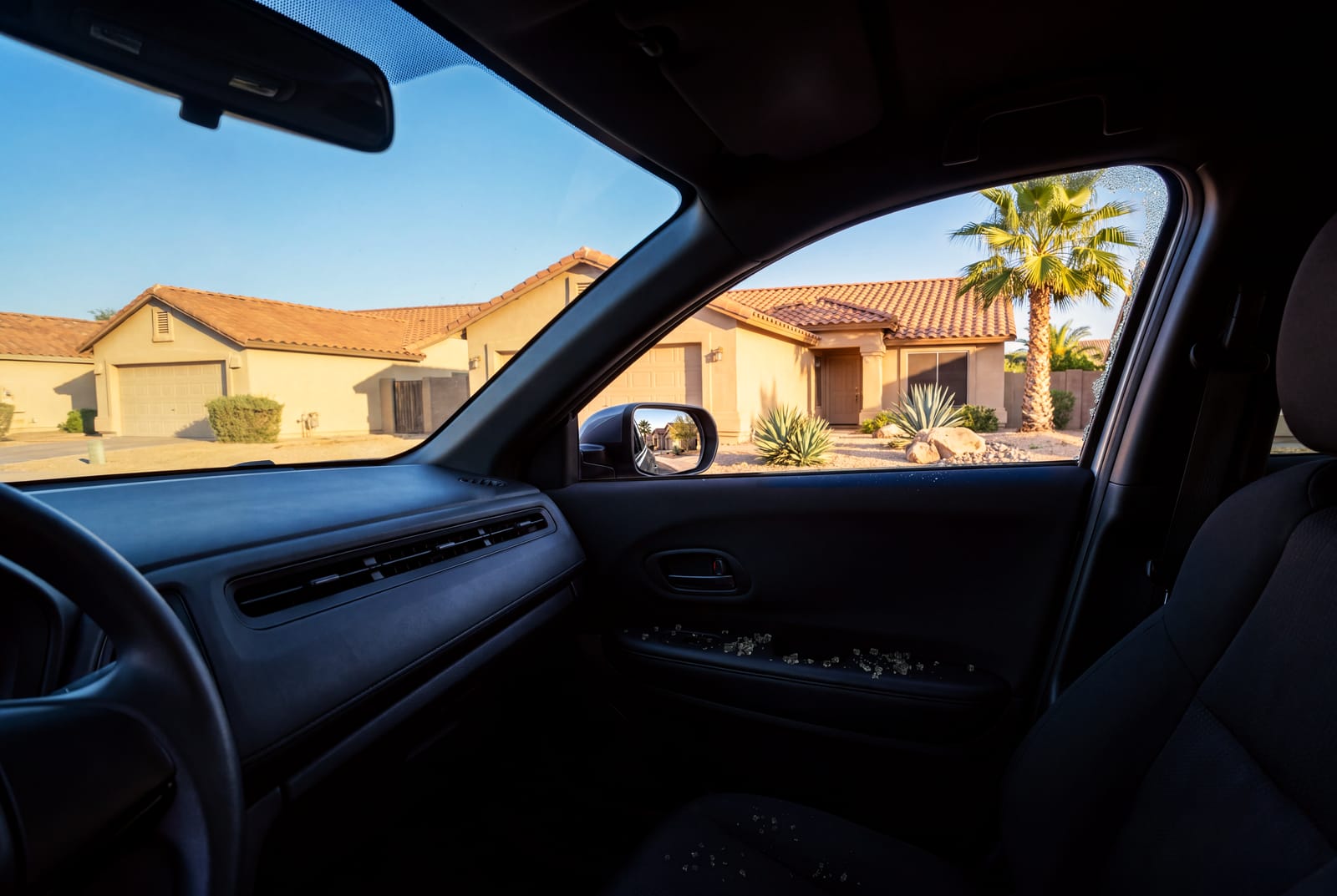 Looking across a car from the driver's seat at the front passenger window, broken out, with a strip of crazed tempered glass left along the top and rear edges of the frame and loose glass pieces on the door trim and the seat, toward a quiet street of stucco homes in the afternoon sun.